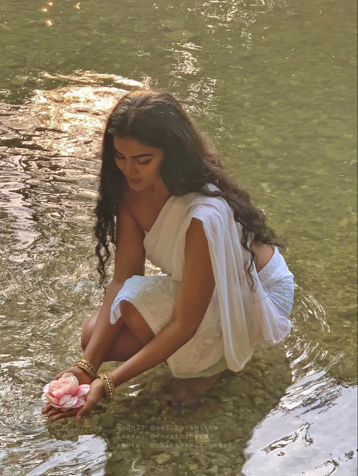 A woman kneeling in a shallow river, offering a rose to the water